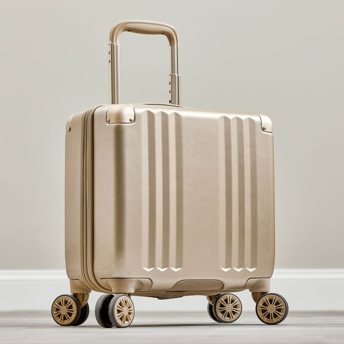 Suitcase stands upright, wheels visible, handle extended; positioned on a wooden floor against a plain, light-colored wall.