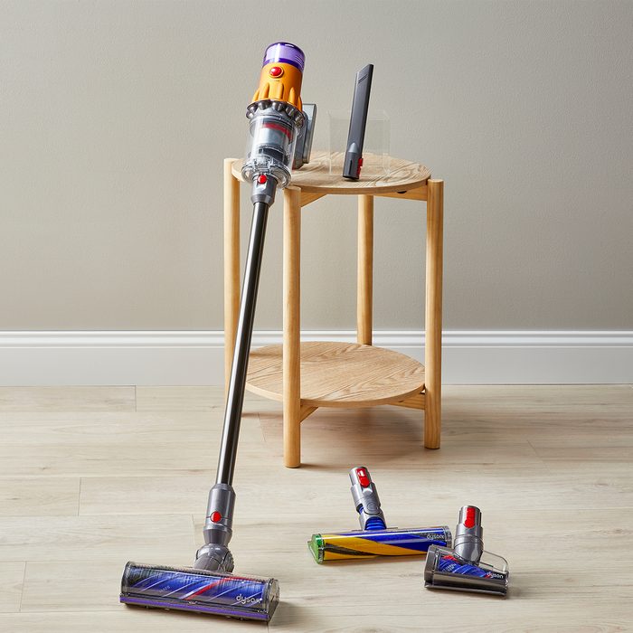 A vacuum cleaner rests against a wooden table on a hardwood floor, with attachments placed nearby.