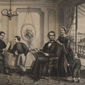 A family sits and stands in a parlor with a portrait. The room has ornate decorations, and a child holds a flag near the window.