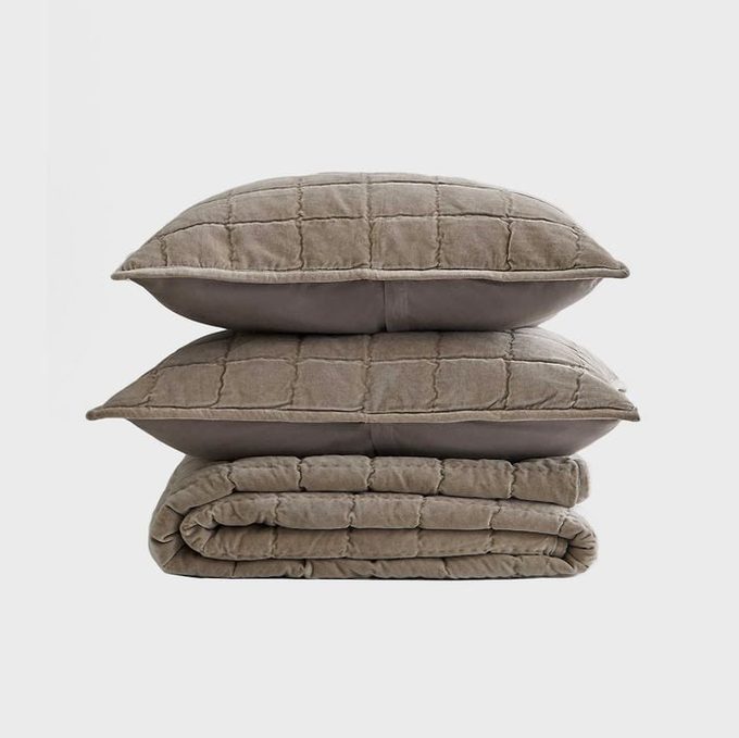 Two beige, quilted pillows stacked on a matching folded blanket, set against a plain white background.