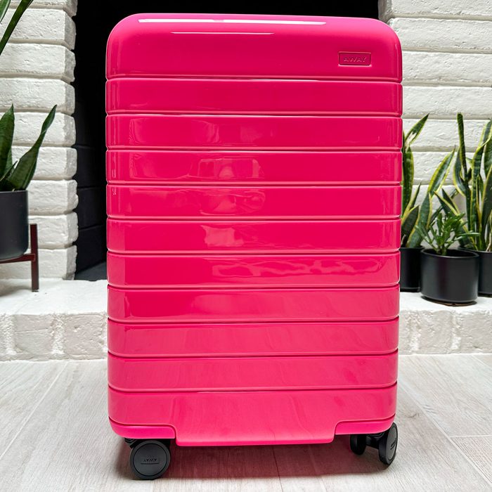 A vibrant pink suitcase rests on four wheels, positioned indoors against a white brick wall, surrounded by several potted plants.