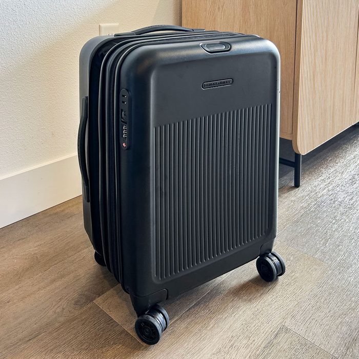 A black rolling suitcase stands upright with four wheels beside a wooden cabinet on a hardwood floor.