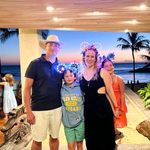 A family poses together, wearing festive headpieces, against a sunset backdrop by the ocean with palm trees and decorative plants.