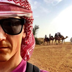 A person with sunglasses and headscarf stands in a desert; camels with riders walk in the background under a blue sky.