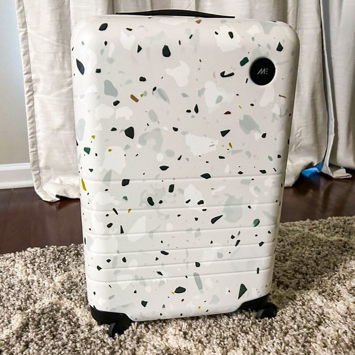 Suitcase stands upright, featuring a terrazzo pattern with black wheels, in a room with wooden flooring and cream curtains.
