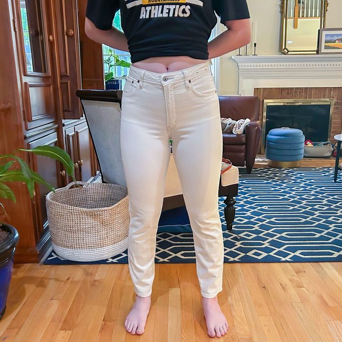 Person wearing white jeans stands in living room, hands behind back, on a patterned rug. Text on shirt: 