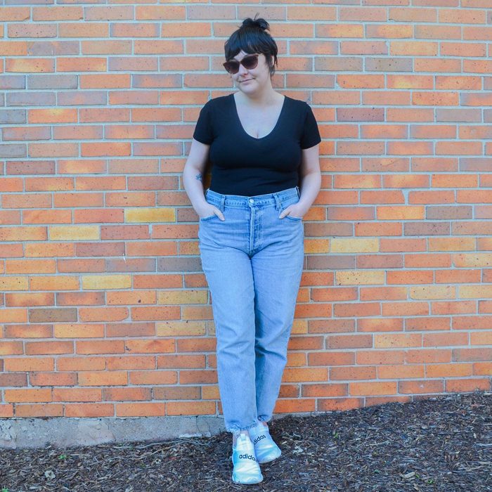 Person stands casually with hands in pockets, wearing sunglasses and leaning against a brick wall outside. Ground is covered with mulch.