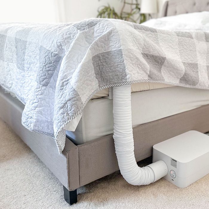 A white vent hose connects a machine to a bed, under a checkered quilt, in a bright, carpeted bedroom.