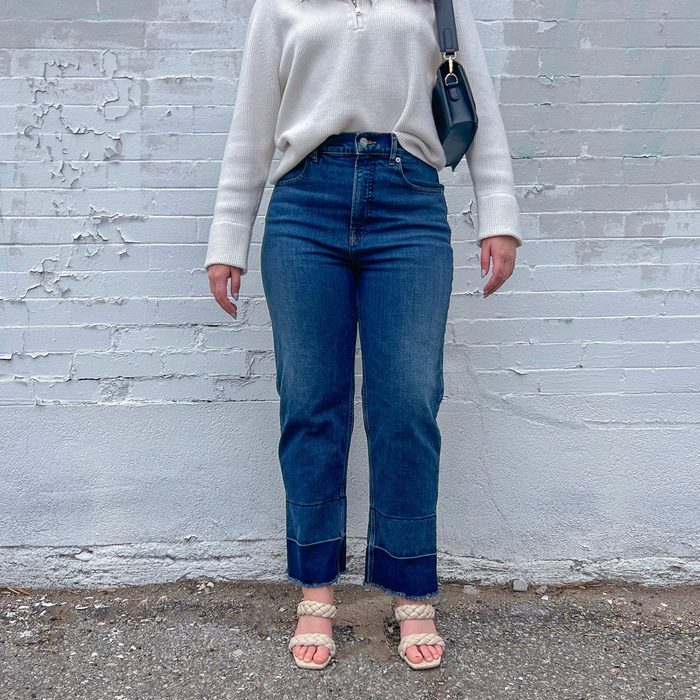 Person standing; wearing a white sweater, blue jeans, and sandals; near a textured, light-colored brick wall.