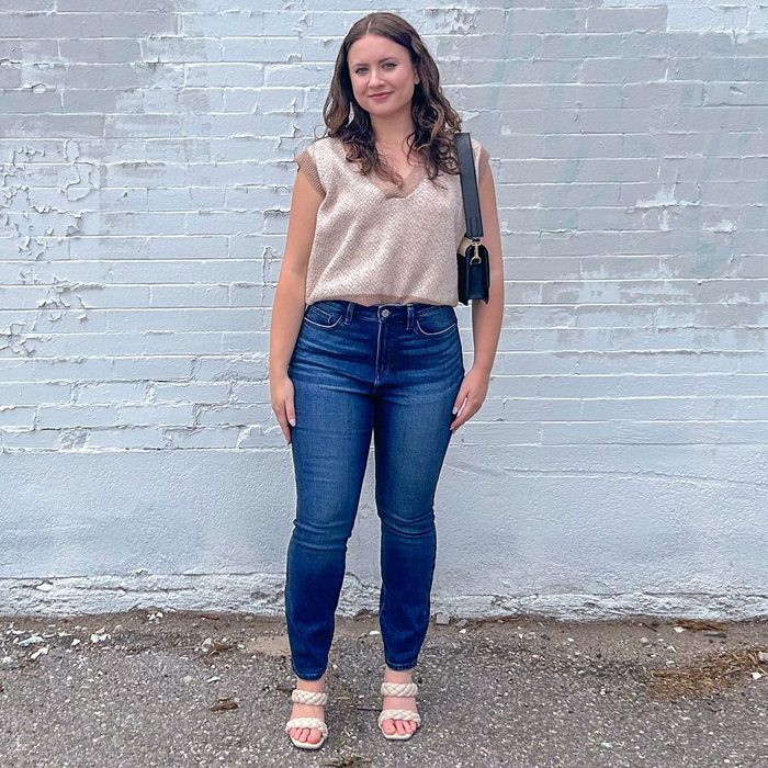 A woman stands smiling, wearing a patterned top and jeans, against a weathered white brick wall.