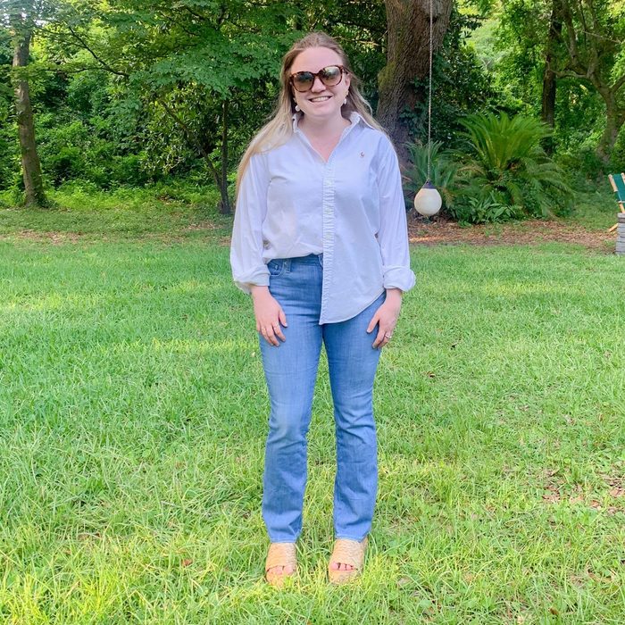 Person standing, smiling, in front of a wooded area wearing sunglasses, a white shirt, and blue jeans on a grassy lawn.