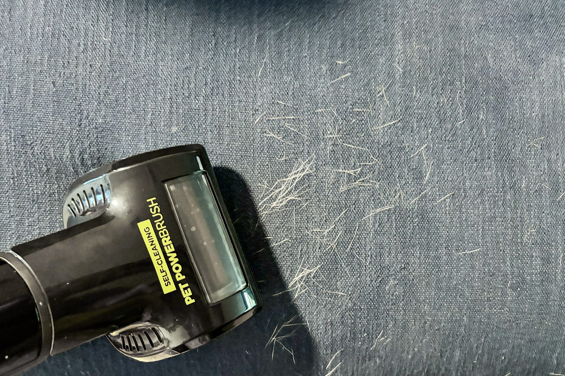 Vacuum powerbrush cleaning pet hair on blue fabric. Text on vacuum reads "Self-Cleaning Pet Powerbrush."