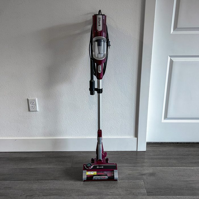 A vacuum cleaner stands upright against a wall on a wooden floor, with cord coiled neatly.