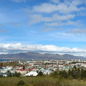 Buildings with colorful roofs sit quietly beneath a partly cloudy sky, surrounded by a mountainous horizon and lush green trees.
