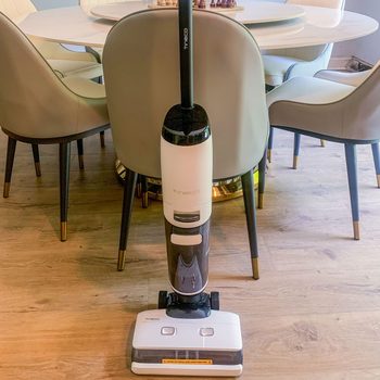 A vacuum cleaner stands idle in a dining room with light wood floors and beige chairs around a circular table.