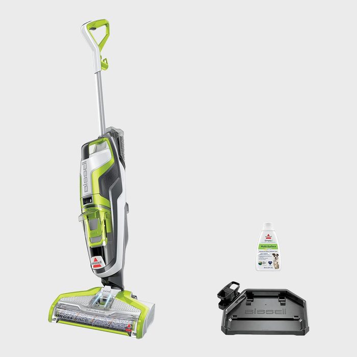 Vacuum cleaner standing upright, accompanied by a cleaning solution bottle and a tray, set against a plain gray background.