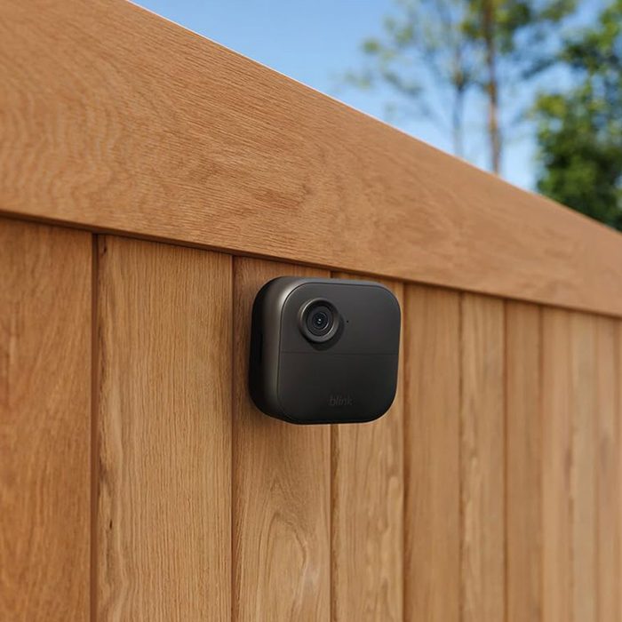 A black security camera is mounted on a wooden fence outdoors, with trees and blue sky in the background.