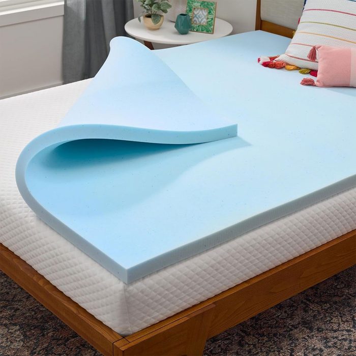 Foam topper lies partially rolled on a bed, atop a white mattress, in a cozy bedroom with pillows and a small table nearby.
