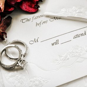 Invitation card with embossed floral design lies beneath two diamond rings, accompanied by text: 
