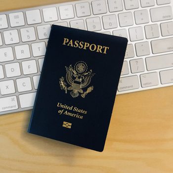 A United States passport lies on a wooden desk, partially covering a white computer keyboard. The passport is closed and its cover, displaying the U.S. emblem and the word "PASSPORT," is clearly visible.
