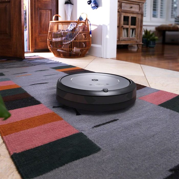 Robot vacuum cleaning a colorful carpet in a modern, well-lit living room with wood and tile flooring, near an open wooden door.