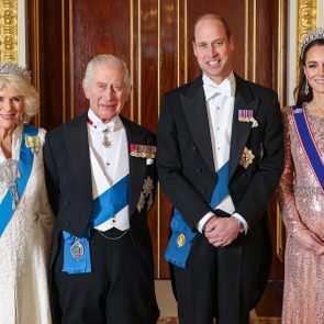 Four elegantly dressed individuals stand smiling, wearing formal attire and sashes, inside an ornately decorated room with gold accents and a polished wooden backdrop.