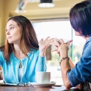 Two women converse at a table; one gestures dismissively while the other listens attentively in a bright cafe setting.