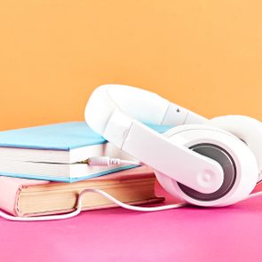 Headphones rest on stacked books, one blue, the other pink; set against an orange background and pink surface.