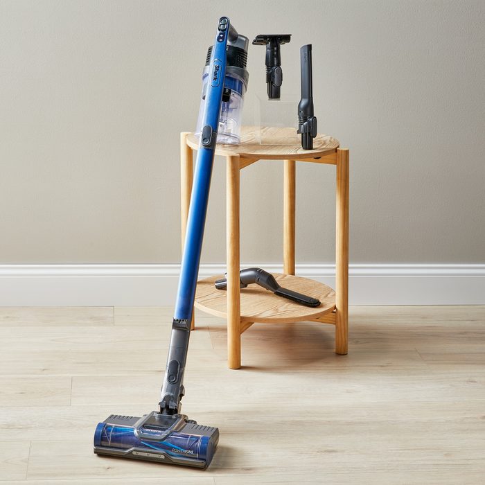 A blue cordless vacuum stands near a small wooden table, surrounded by various attachments on and above the table, in a bright room.