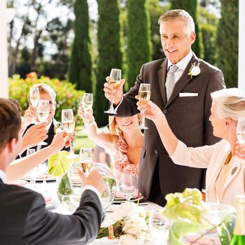 Man raises glass in celebratory toast; others join around an outdoor dining table with floral centerpieces, dressed formally in bright sunlight.