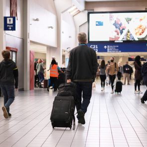 Traveler pulls suitcase through bustling airport terminal with overhead signs and people moving in various directions.