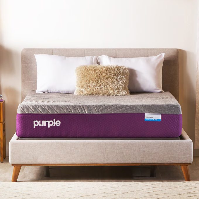 Mattress sits on a wooden bed frame, featuring a purple cover. It is flanked by white pillows and a fuzzy cushion in a bedroom.