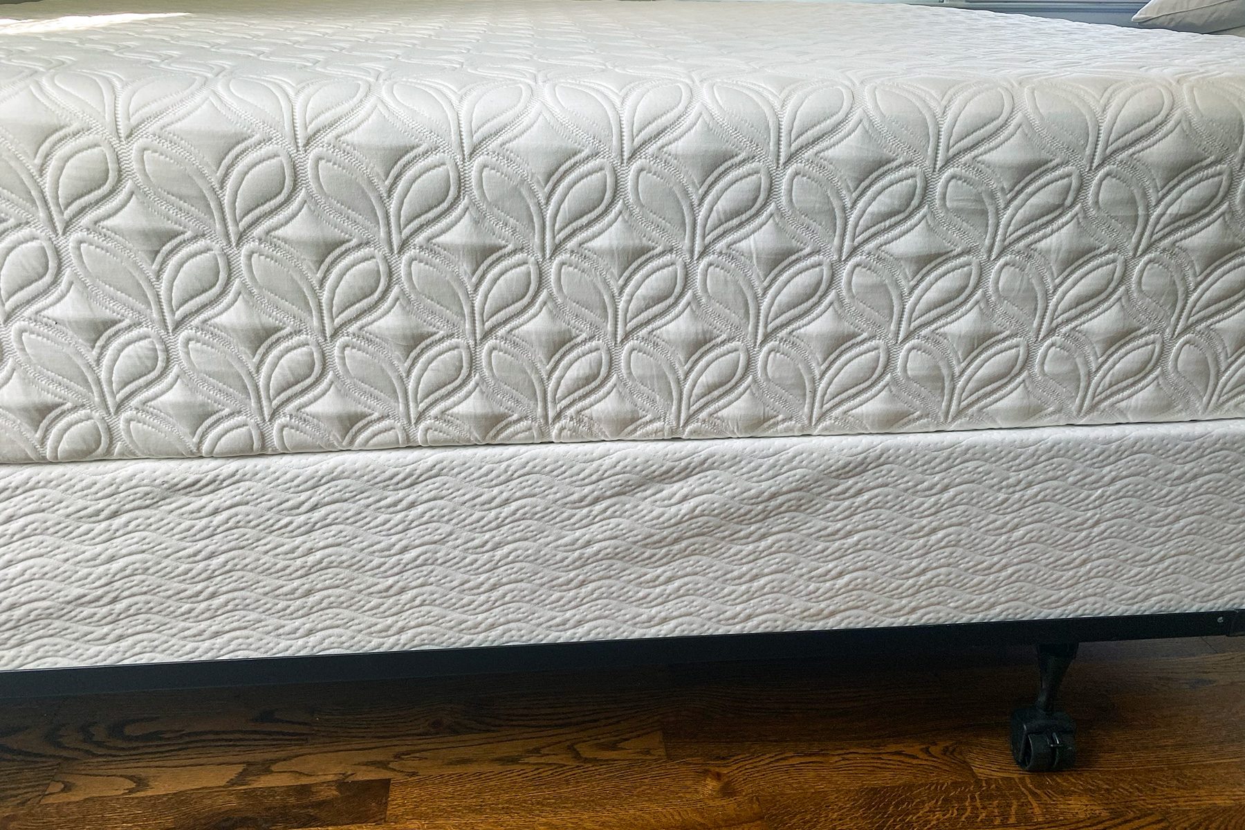 Mattress features embossed leaf pattern, resting on a bed frame with caster wheels, situated on wooden flooring.