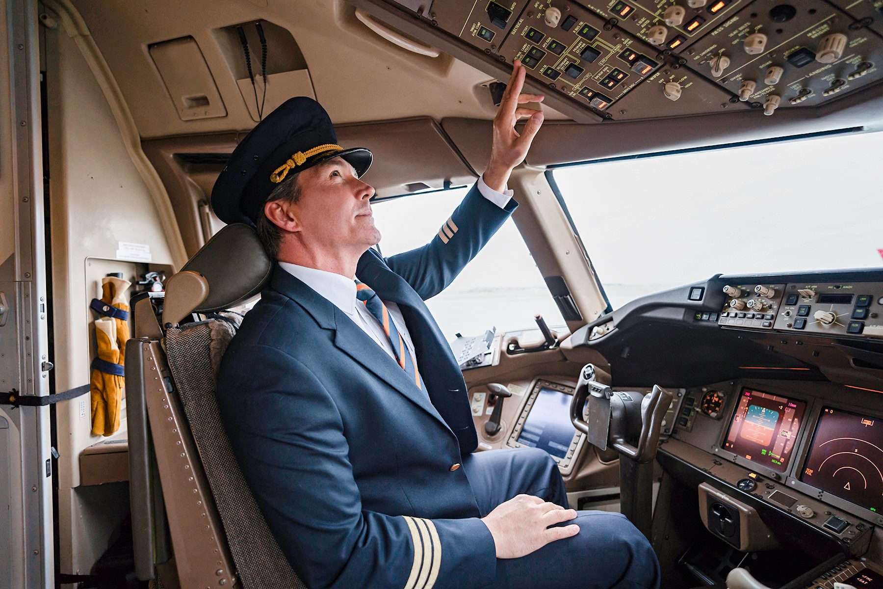 Pilot adjusts controls overhead in airplane cockpit, surrounded by various flight instruments and monitors.