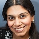 A person smiles warmly while facing forward, wearing hoop earrings and a patterned top, against a plain background.