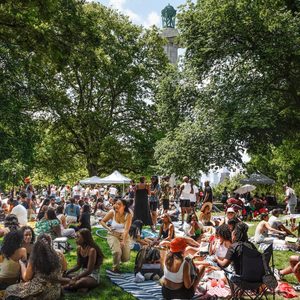 People sitting and socializing on blankets in a park, surrounded by trees and tents, with a distant tower visible through the canopy.
