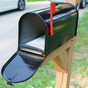 Mailbox stands open with a red flag raised, on a wooden post beside a driveway with a parked white car.