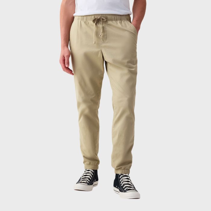 Beige jogger pants are worn by a standing person with hands in pockets, wearing black sneakers. The background is plain white.