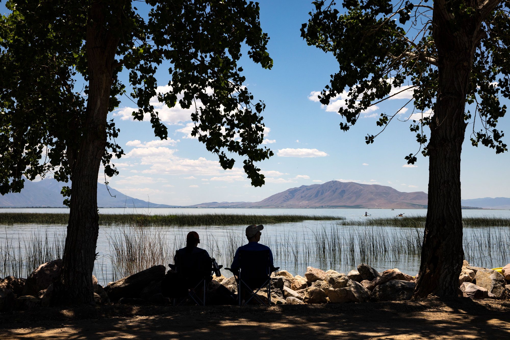 Two people sit on chairs, facing a lake bordered by rocks and tall grass, under leafy trees with mountain and sky in the background.