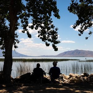 Two people sit on chairs, overlooking a lake with reeds, beneath tree branches, mountains visible in the background under a partly cloudy sky.