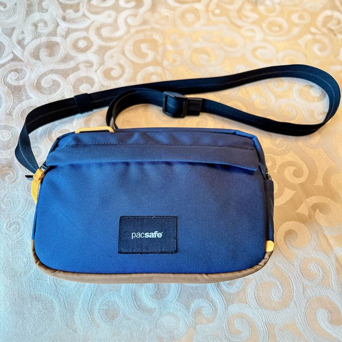 A blue Pacsafe bag rests on a swirly-patterned fabric, featuring a black strap and yellow zipper pull.