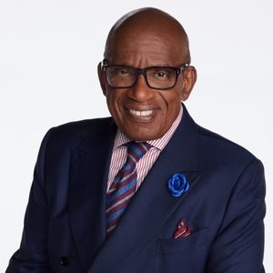 Man smiling, wearing glasses, a navy suit, and a striped tie, with a blue flower lapel pin, against a plain light background.