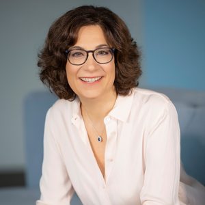 A woman smiles, wearing glasses and a white blouse, in a softly lit room with a blurred blue background.