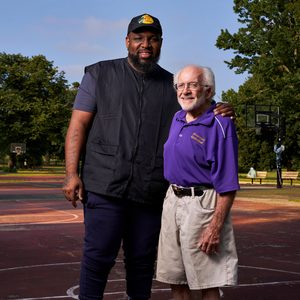 Two men stand together smiling on a basketball court, surrounded by trees. One wears a black vest; the other, a purple shirt with 