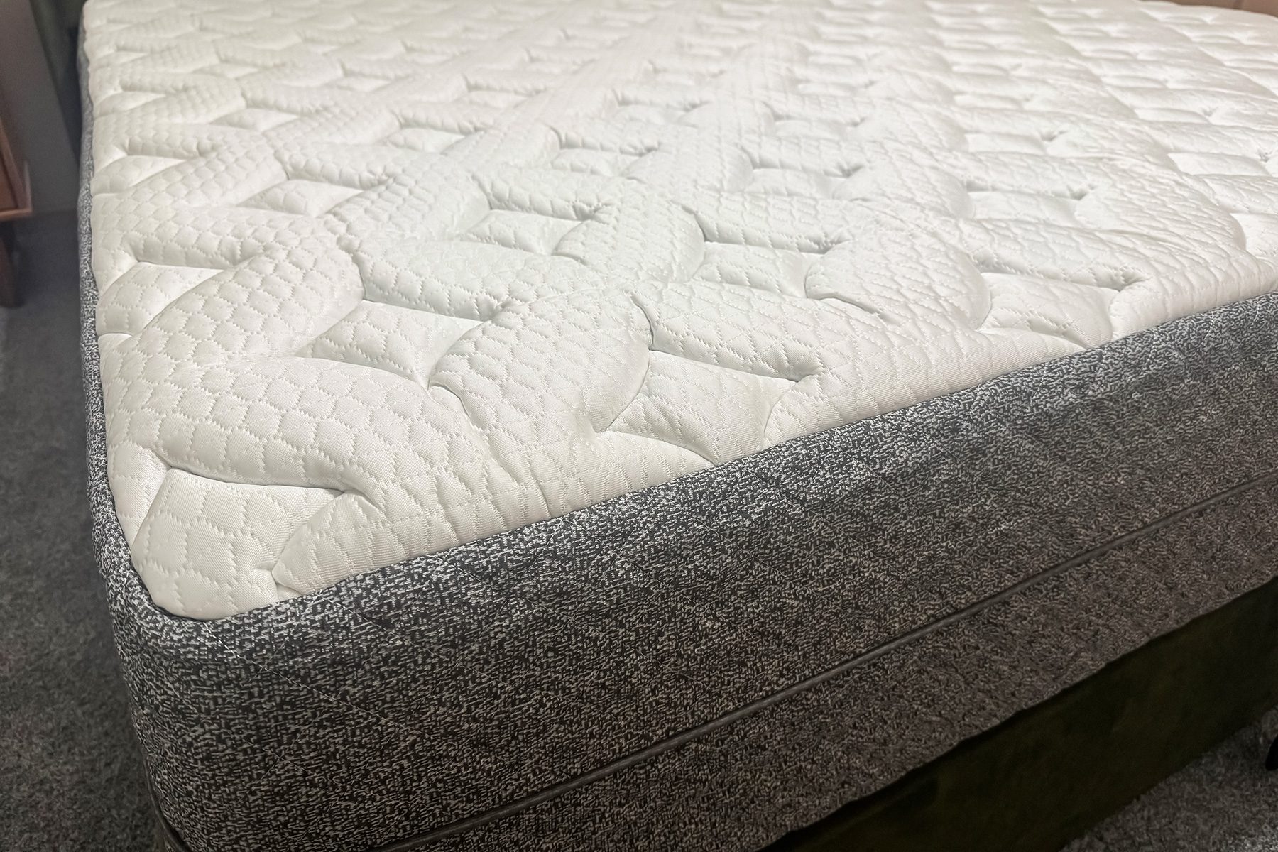 A mattress with a quilted white surface and textured gray sides rests on a carpeted floor in a room corner.