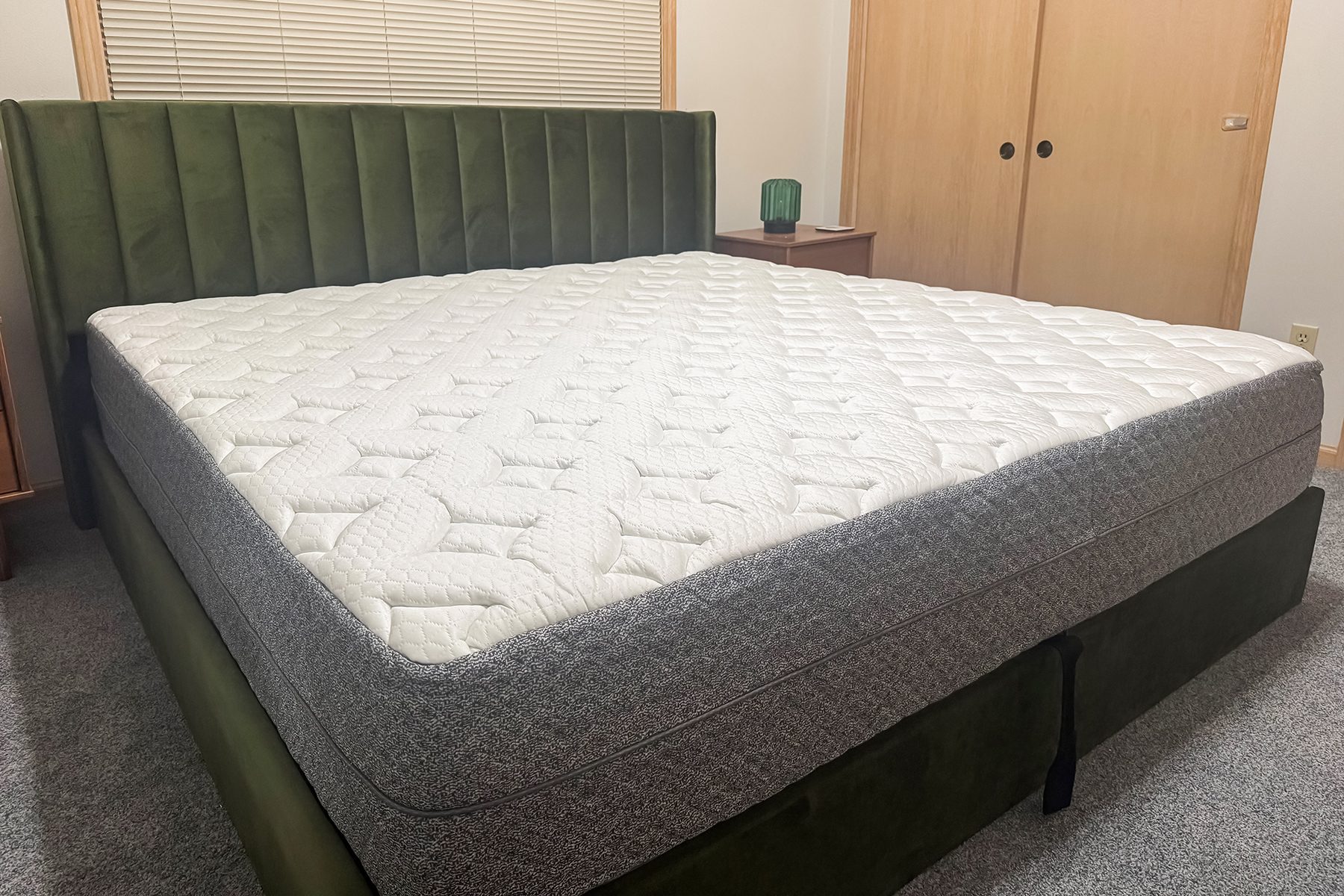 Bed features a large, quilted mattress on a green upholstered frame, situated in a bedroom with carpet, nightstand, and closed wooden closet doors.