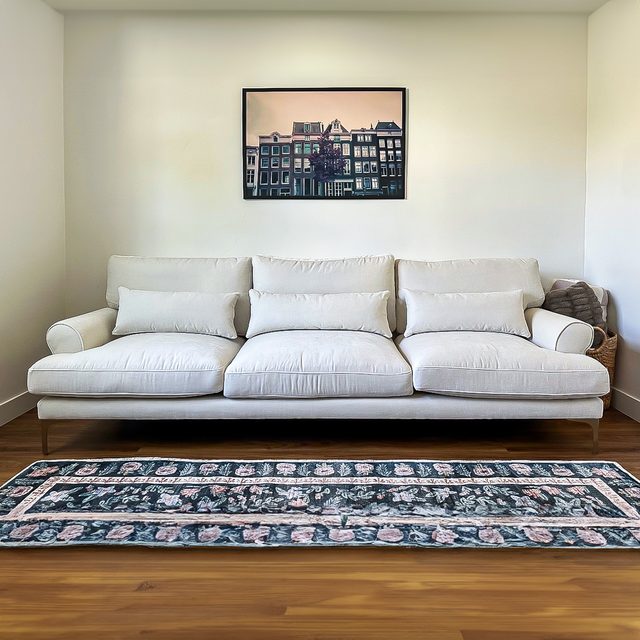White sofa sits quietly against a wall with framed urban photo above; patterned rug lays on wooden floor in a minimalist room.