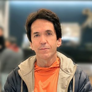 A man stands calmly, wearing an orange shirt and gray jacket, with a blurred indoor background suggesting a casual setting.