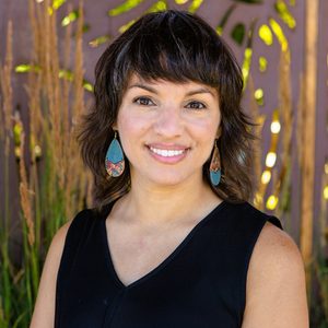 Person smiling, standing still, wearing colorful earrings and a black top; background shows tall grasses and a blurred, patterned wall.