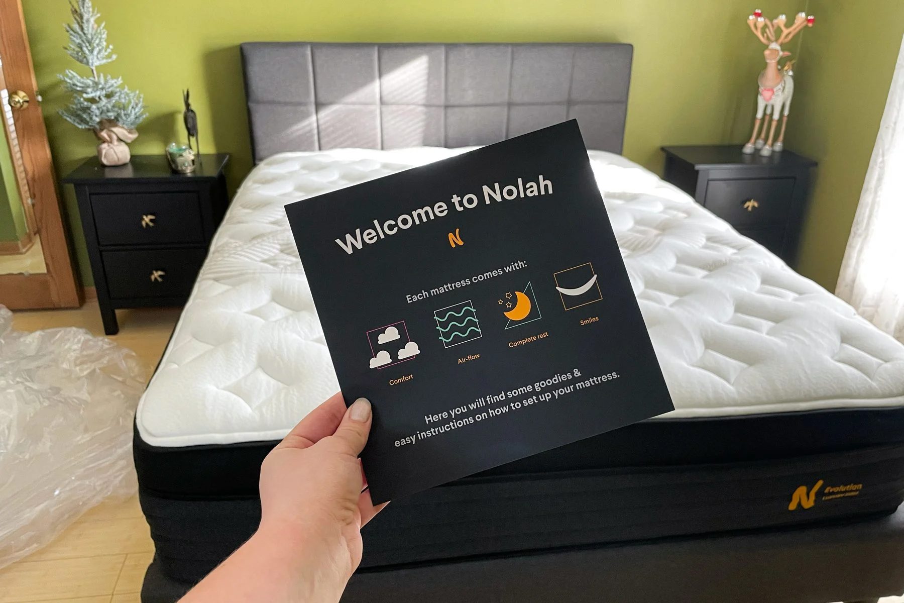 Hand holds a guide, welcoming to Nolah. Displaying features: comfort, airflow, restful sleep, smiles. Context: bedroom with bed, pillows, and side tables.